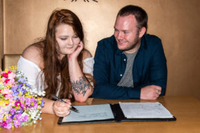Martha and Andrew smiling while Martha signs a the wedding register at a table next to a bouquet of colorful flowers.