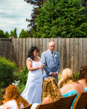 Bride in a white lace wedding dress reading vows to the groom in a light blue suit during an outdoor garden ceremony.