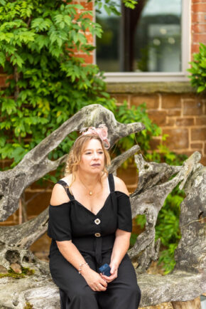 A moment of contemplation for the mother of the bride - Oak Tree Barns, Narborough 2021 Mother of the bride taking a quiet moment to breathe, sitting on a wooden bench in a black outfit and pink fascinator.