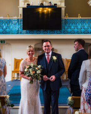 Bride Pamela holding her bouquet and groom Philip standing together at the front of the venue during their wedding ceremony.