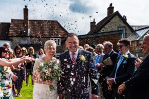 Bride and groom Pamela and Philip laughing as wedding guests throw colorful confetti during their outdoor reception.