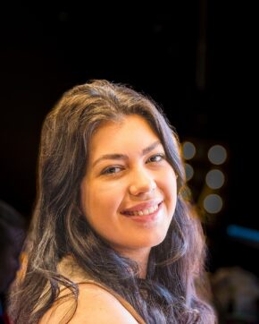 Backstage Portrait from 'Miss Caradog is a Serial Killer' Close-up portrait of a smiling young woman with dark hair against a dark background with soft bokeh lights.