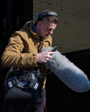 Behind-the-scenes shot on the set of Miss Caradog is a Serial Killer Candid portrait of a sound recordist using a hairbrush to groom a furry microphone deadcat on a film set.