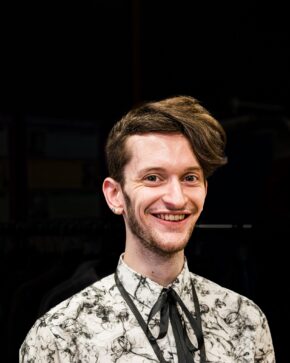 Backstage Portrait from 'Miss Caradog is a Serial Killer' Portrait of a stylish young man wearing a patterned shirt and black bow tie against a dark background.