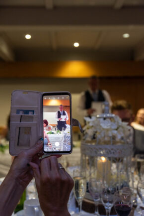 A wedding guest holding a smartphone in a wallet case to record or photograph a wedding speech at the reception.