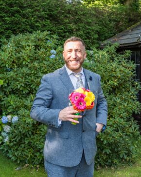 Guest smiling happily in a blue textured suit while playfully holding a vibrant bouquet of pink and yellow gerbera daisies.