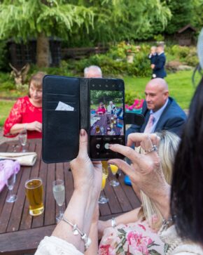 Close-up of a wedding guest using a smartphone in a black wallet case to take a picture of people sitting at an outdoor garden table reception.