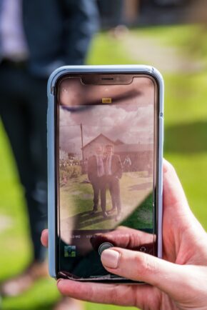 A wedding guest holding a smartphone and taking a photo of two people outside during a wedding reception.