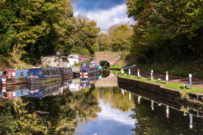 Fine art canal photography by Charlie Hannah ARPS, featuring a quiet canal lined with colourful moored narrowboats beneath a stone bridge framed by autumn trees.