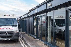 Exterior view of a gray commercial building with a white delivery van parked alongside it while the driver has a conversation with the business owner.
