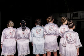 A candid documentary-style photograph showing the bride and bridal party lineup from behind, wearing matching pink satin robes with custom floral embroidery.