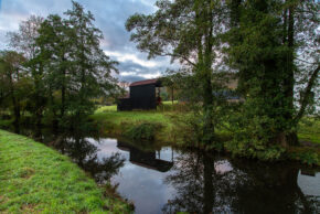 A black wooden shed and lush green trees reflecting perfectly on the calm water of a UK canal under a cloudy sky.