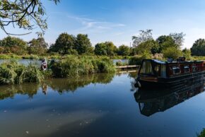 Fine art canal photography by Charlie Hannah ARPS, showing a lone angler fishing on a tranquil canal bank alongside a colourful moored narrowboat reflecting in the calm water.