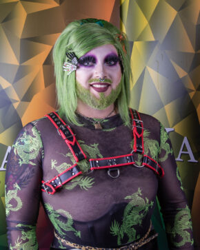 An expressive alternative portrait featuring a drag performer with green hair and a beard, styled with a red leather harness and bold purple eyeshadow. Close-up portrait of a drag performer with green hair, a beard, and a red leather harness over a sheer patterned top.