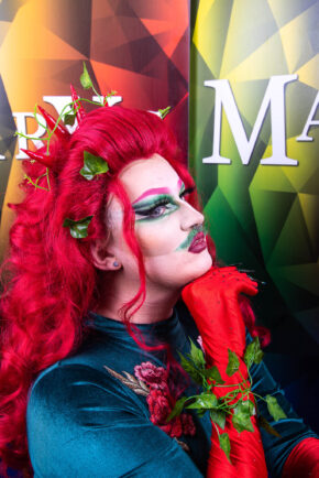Alternative formal portrait capturing a drag artist in a rich green velvet top, bold red hair accented with ivy, and striking theatrical makeup. Striking portrait of a drag artist wearing a green velvet outfit, red hair with ivy accents, and red gloves.