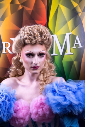 A stunning, relaxed formal portrait of a Drag Race star featuring dramatic makeup, classic vintage hair styling, and vibrant blue and pink tulle fashion. Text: High-fashion portrait of a former RuPaul's Drag Race contestant styled in elaborate blue and pink tulle with vintage curls.