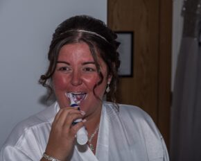 A humorous, relaxed shot of a bridesmaid getting ready and brushing her teeth in her bridal party robe. Bride laughing while brushing her teeth with an electric toothbrush in a white robe on the wedding morning.