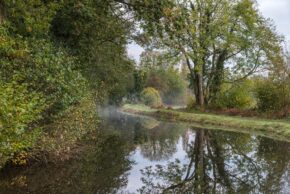 Fine art canal photography by Charlie Hannah ARPS, capturing a misty morning scene with lush autumnal trees and foliage mirrored perfectly in a calm, winding canal.