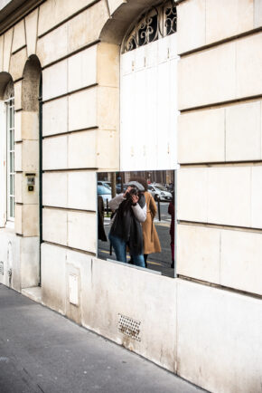 A photographer taking a picture into a wall-mounted mirror that reflects a European street scene.