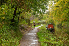 A lone runner jogging along a wet, leaf-strewn canal towpath flanked by autumn trees and a moored narrowboat.