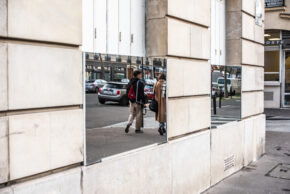 Stone building exterior featuring a mounted mirror that reflects pedestrians walking down a Parisian street.