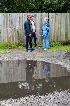 Wide angle view of television cast members standing outdoors near a wooden barrier with a sharp reflection in a puddle.
