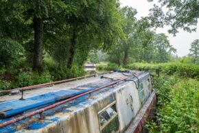 Fine art canal photography by Charlie Hannah ARPS, capturing the weathered blue and red roof of a moored narrowboat resting quietly among overgrown riverside reeds and trees.