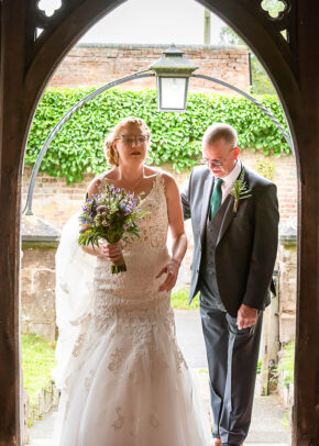 Bride carrying a bouquet walking through an arched doorway with her Dad at the Gemma and Martin wedding, photographed by South Wales wedding photographer Charlie Hannah