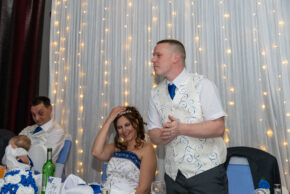 Groom delivering a speech at the head table while the smiling bride adjusts her tiara during a wedding reception, photographed by Charlie Hannah at The Belfry, Nottingham